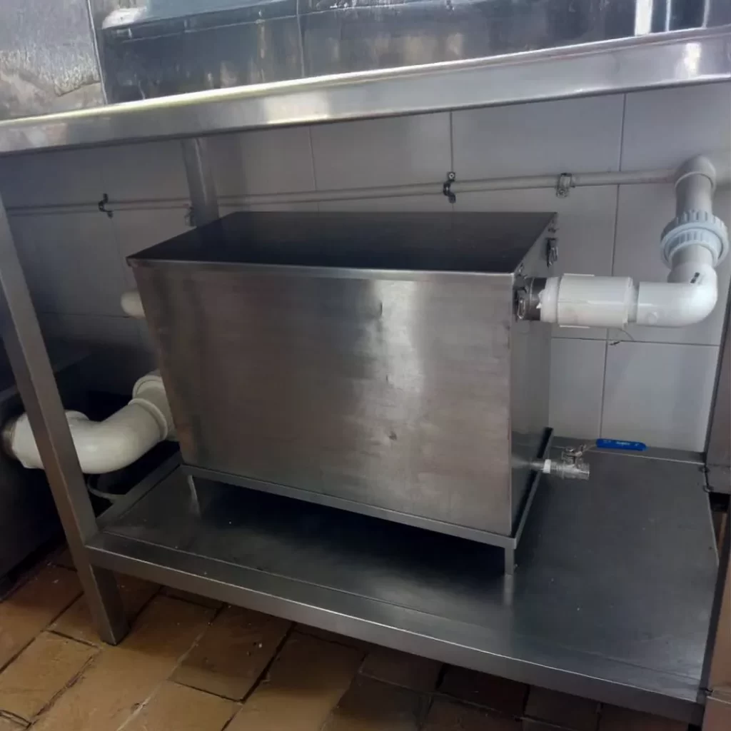 Trampa de grasa en acero inoxidable instalada bajo una mesa de trabajo en cocina industrial, conectada a tubería de PVC.